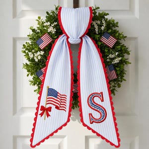 May include: A patriotic wreath with a green base, adorned with small American flags and a blue and white striped sash. The sash features a red scalloped edge, an embroidered American flag, and the letter "S".