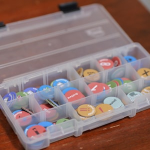 May include: A clear plastic storage container with a hinged lid, featuring multiple internal compartments filled with assorted circular game tokens in various colors including red, blue, green, and tan, some marked with text like Active and Voted.