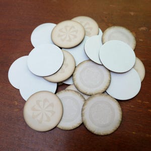 May include: A collection of circular discs in a pile. Some discs are plain white, while others feature a light beige tone with a subtle, darker brown pinwheel or circular pattern printed on the surface. The discs have a smooth, flat appearance.