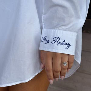 Könnte beinhalten: Weißes Button-Down-Hemd mit marineblauem, gesticktem Text "Mrs. Radivoj" am Ärmel. Ein goldener Ring mit Diamant ist an der Hand zu sehen.