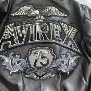 Peut inclure: Veste en cuir noir avec d&eacute;tails brod&eacute;s. Comprend le mot "AVIREX" en grandes lettres, un embl&egrave;me d'aigle et un &eacute;cusson avec le chiffre "75" et le texte "AVIATOR'S KING".