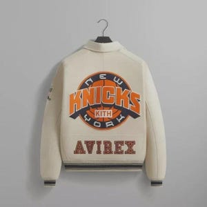 May include: Cream-colored varsity jacket with the New York Knicks logo in orange and blue on the back. The jacket features the "KITH" and "AVIREX" brand names. The jacket has a black and blue striped hem and cuffs.