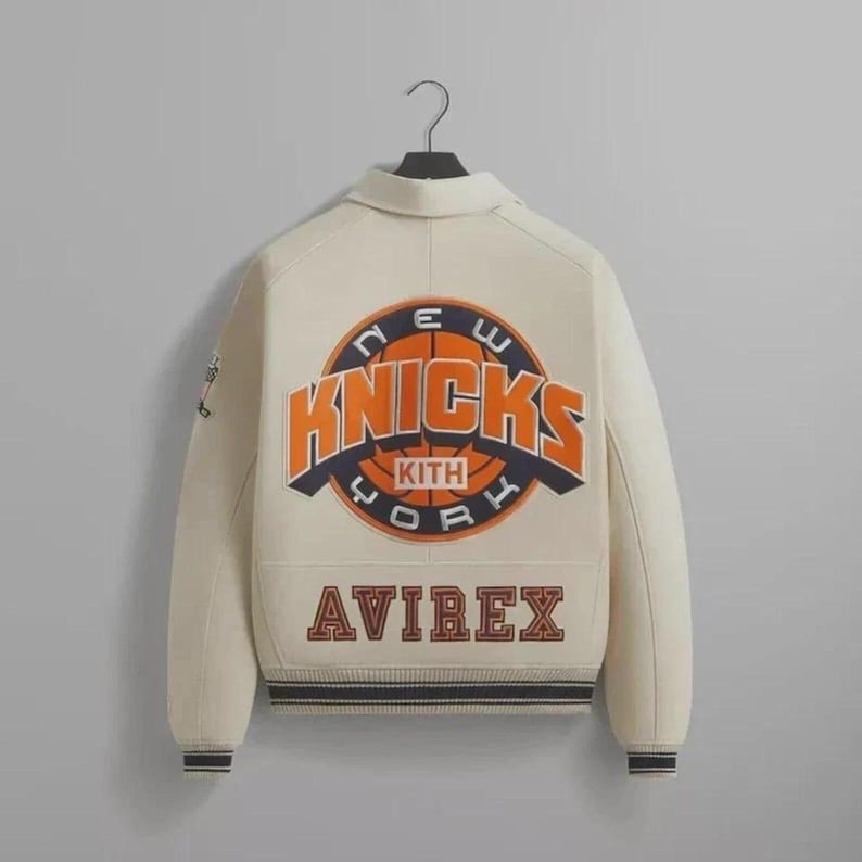 May include: Cream-colored varsity jacket with the New York Knicks logo in orange and blue on the back. The jacket features the "KITH" and "AVIREX" brand names. The jacket has a black and blue striped hem and cuffs.