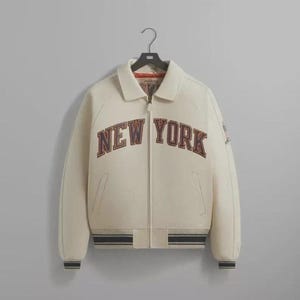 May include: Cream-colored varsity jacket with the text "NEW YORK" in a dark brown and orange color. The jacket has a zip-up front, side pockets, and a ribbed hem and cuffs with black and white stripes. The jacket is hanging on a black hanger.