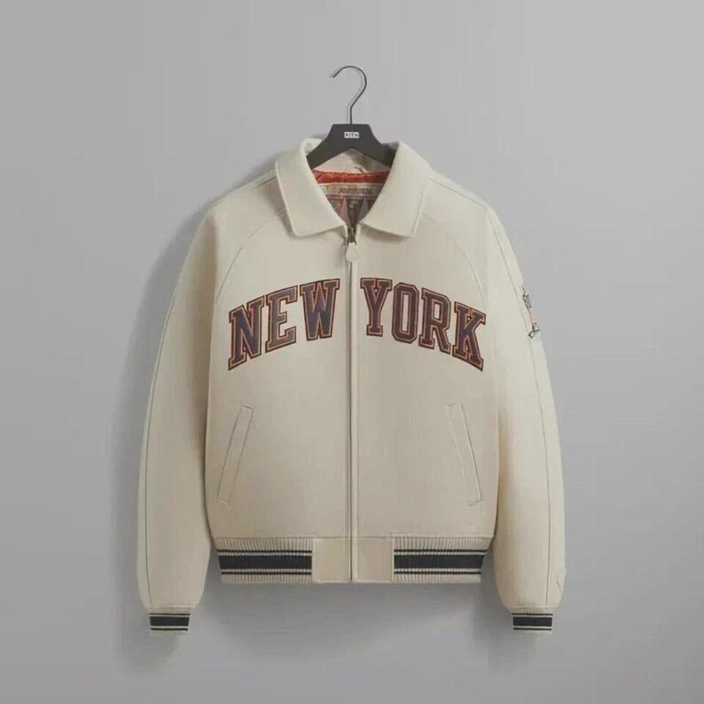 May include: Cream-colored varsity jacket with the text "NEW YORK" in a dark brown and orange color. The jacket has a zip-up front, side pockets, and a ribbed hem and cuffs with black and white stripes. The jacket is hanging on a black hanger.