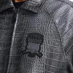Pode incluir: Casaco cinzento texturizado com padr&atilde;o de pele de crocodilo. O casaco tem fecho de correr e gola. Um patch no peito diz "AVIREX NYC ESTABLISHED 1975" com um desenho de bandeira.