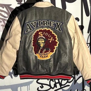 May include: A black and beige leather jacket featuring the word "AVIREX" on the back. The jacket has a patch with a Native American portrait and the words "SIOUX FOOTBALL TEAM". The cuffs are red and white striped.