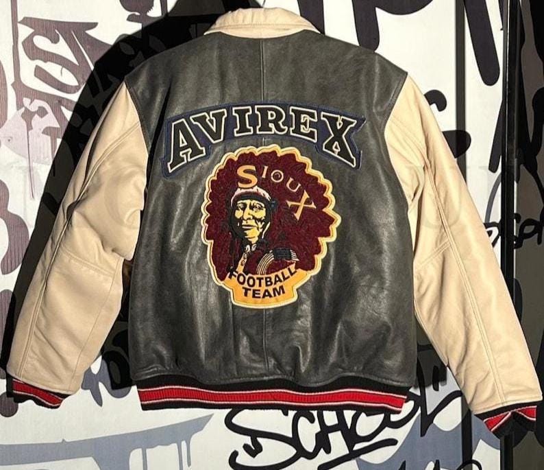 May include: A black and beige leather jacket featuring the word "AVIREX" on the back. The jacket has a patch with a Native American portrait and the words "SIOUX FOOTBALL TEAM". The cuffs are red and white striped.