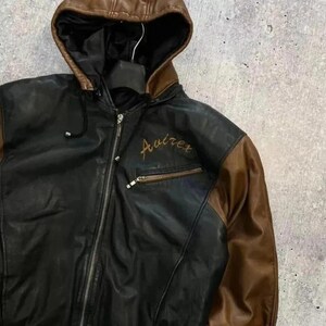 Puede incluir: Una chaqueta con capucha de cuero negro y marr&oacute;n que presenta una cremallera frontal completa, una capucha con cord&oacute;n y un bolsillo en el pecho con cremallera. La palabra Avirex est&aacute; bordada en hilo dorado en el pecho izquierdo del cuero.
