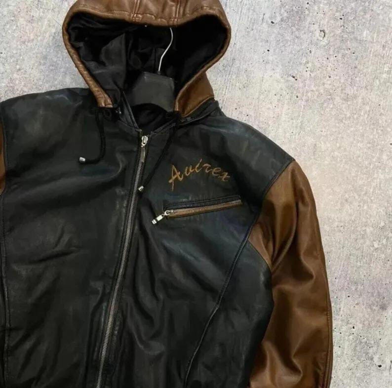 Puede incluir: Una chaqueta con capucha de cuero negro y marr&oacute;n que presenta una cremallera frontal completa, una capucha con cord&oacute;n y un bolsillo en el pecho con cremallera. La palabra Avirex est&aacute; bordada en hilo dorado en el pecho izquierdo del cuero.