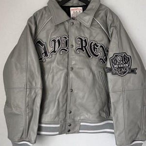 May include: Gray leather jacket with the word "AVIREX" in large black lettering across the chest. The jacket has a button-up front, white trim, and a patch that says "AVIREX PALACE". The jacket is hanging on a black hanger.