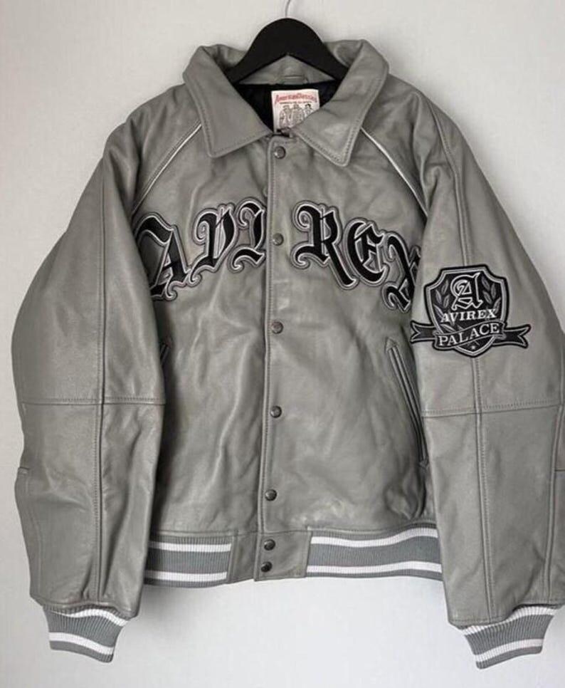 May include: Gray leather jacket with the word "AVIREX" in large black lettering across the chest. The jacket has a button-up front, white trim, and a patch that says "AVIREX PALACE". The jacket is hanging on a black hanger.