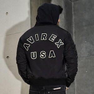 May include: Black bomber jacket with the text "AVIREX USA" in white block letters on the back. The jacket has a hood and ribbed cuffs. The model is wearing dark jeans.