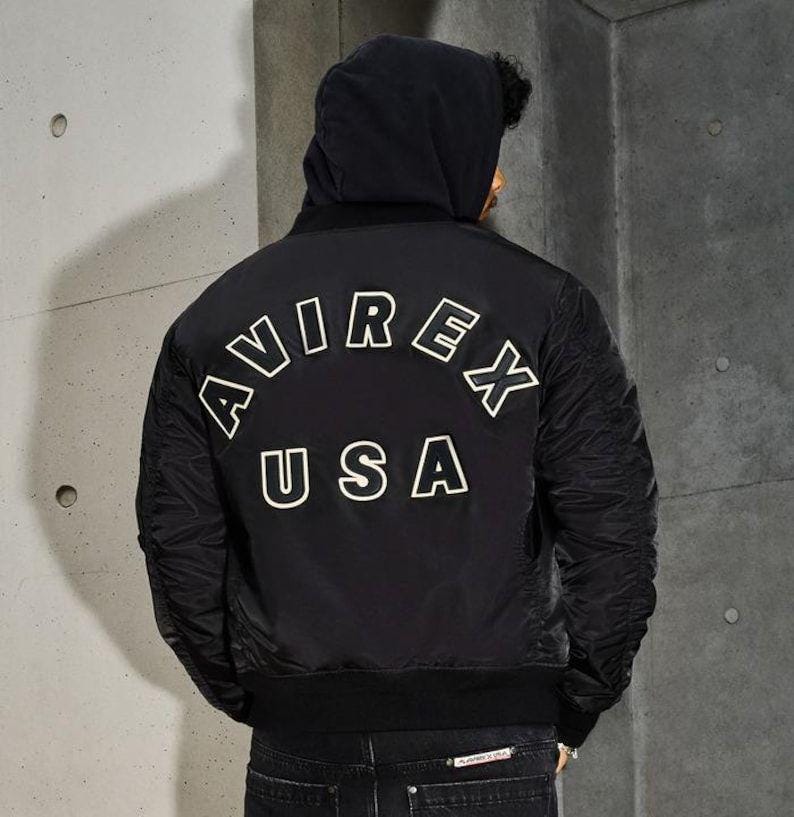 May include: Black bomber jacket with the text "AVIREX USA" in white block letters on the back. The jacket has a hood and ribbed cuffs. The model is wearing dark jeans.