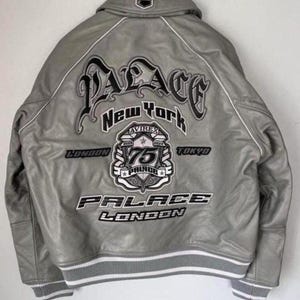 May include: Gray leather jacket with white trim and embroidered logos. The back features the word "PALACE" in large, stylized lettering, along with "New York", "London", and "Tokyo". A patch reads "75 PALACE".