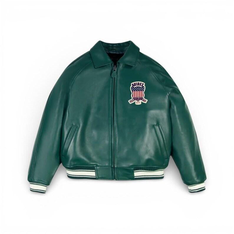 Peut inclure: Veste de bombardier en cuir vert avec fermeture &eacute;clair, col et poignets et ourlet c&ocirc;tel&eacute;s. La veste comporte un &eacute;cusson sur la poitrine avec le texte "AVIREX" et un motif incorporant le drapeau am&eacute;ricain.