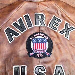 May include: Brown leather jacket with the word "AVIREX" in black and white lettering across the back. A circular patch reads "ALL AMERICANS ESTABLISHED 1975 NY" with a red, white, and blue shield. The word "USA" is also displayed.
