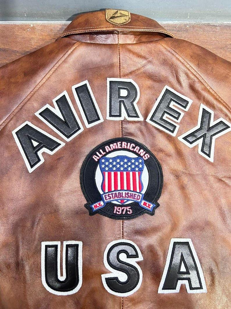 May include: Brown leather jacket with the word "AVIREX" in black and white lettering across the back. A circular patch reads "ALL AMERICANS ESTABLISHED 1975 NY" with a red, white, and blue shield. The word "USA" is also displayed.