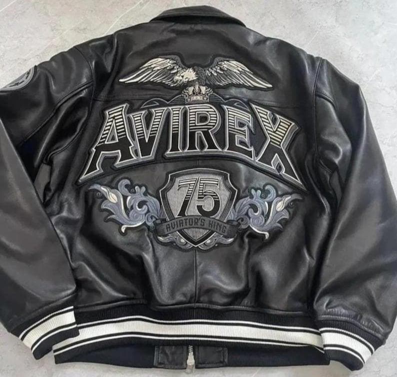 Peut inclure: Veste en cuir noir avec le logo Avirex et un grand embl&egrave;me d'aigle au dos. La veste pr&eacute;sente un ourlet c&ocirc;tel&eacute; avec des rayures blanches et une fermeture &eacute;clair. Le design comprend le texte "75 Aviator's King".