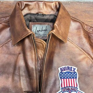 May include: Brown leather jacket with a collar and a zipper closure. Features a patch with the text "AVIREX ESTABLISHED 1975 N.Y." on the front. The jacket has a "Lamhah" label.