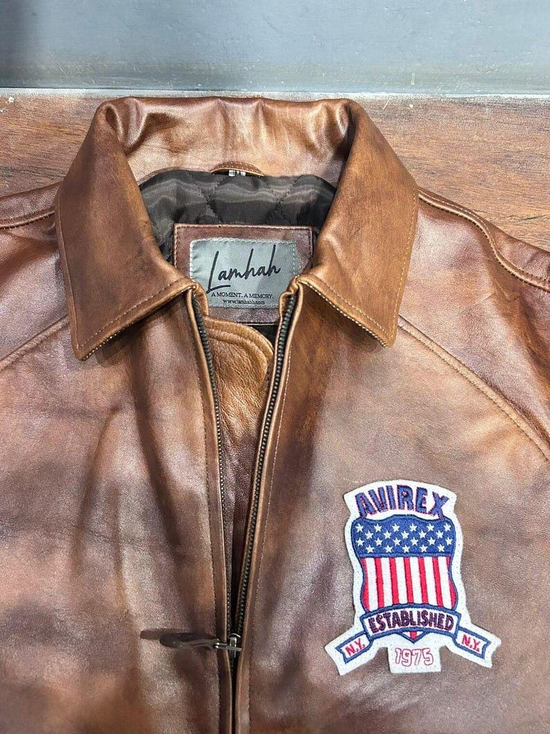 May include: Brown leather jacket with a collar and a zipper closure. Features a patch with the text "AVIREX ESTABLISHED 1975 N.Y." on the front. The jacket has a "Lamhah" label.