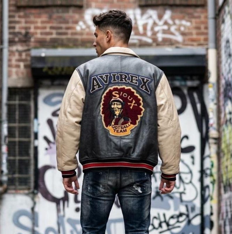May include: Black leather jacket with beige sleeves and a cream collar. The back displays an "AVIREX" patch above a Sioux football team logo. Red and black striped trim at the waist and cuffs.