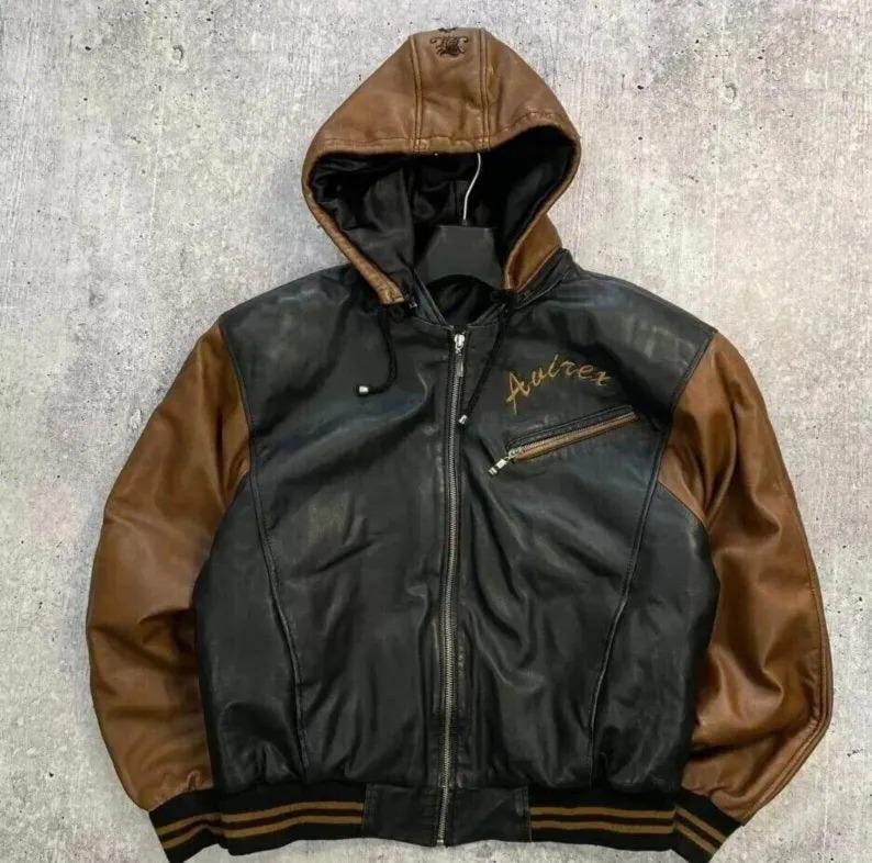 Puede incluir: Una chaqueta con capucha de dos tonos que presenta un cuerpo de cuero negro y mangas de cuero marr&oacute;n. La chaqueta incluye un cierre de cremallera frontal, un bolsillo en el pecho con cremallera y el texto bordado Avirex, y pu&ntilde;os y dobladillo acanalados.