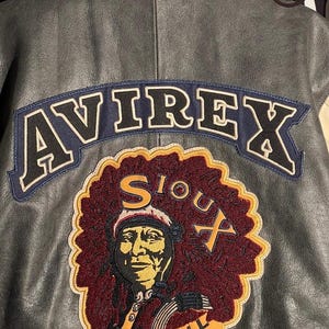 May include: A grey leather jacket featuring the word "AVIREX" in white outlined letters on a navy blue patch. Below is a patch with a Native American portrait and the words "SIOUX FOOTBALL TEAM" in yellow.