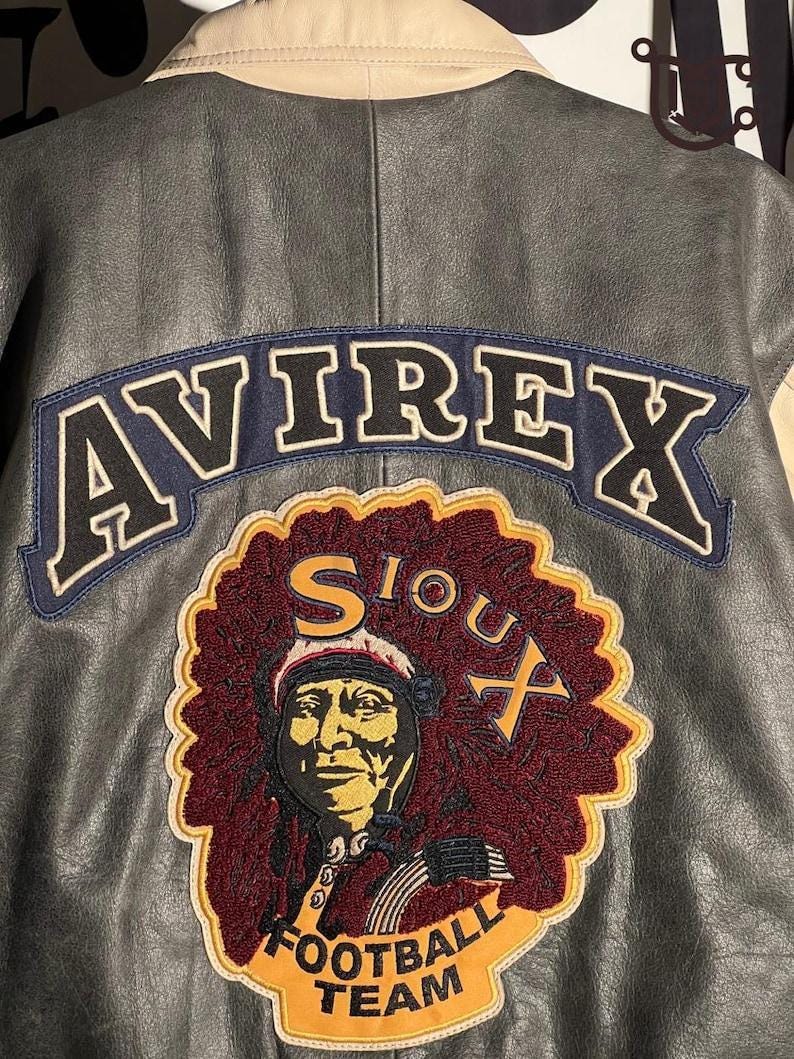 May include: A grey leather jacket featuring the word "AVIREX" in white outlined letters on a navy blue patch. Below is a patch with a Native American portrait and the words "SIOUX FOOTBALL TEAM" in yellow.