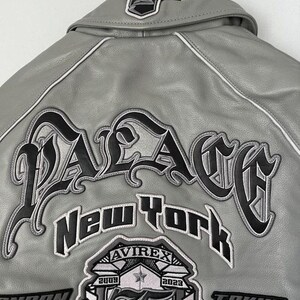 May include: A gray leather jacket with white trim and black embroidered lettering. The back of the jacket features the word "PALACE" in large, stylized letters, along with "New York" and other patches.