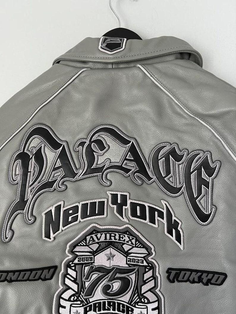 May include: A gray leather jacket with white trim and black embroidered lettering. The back of the jacket features the word "PALACE" in large, stylized letters, along with "New York" and other patches.