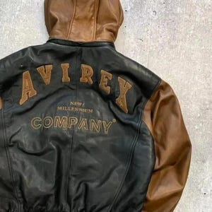 Puede incluir: Una chaqueta de cuero negra y marr&oacute;n con capucha de cuero marr&oacute;n. La parte posterior muestra el texto bordado AVIREX en letras grandes de color marr&oacute;n, con el texto NEW MILLENNIUM COMPANY bordado en letras doradas m&aacute;s peque&ntilde;as debajo.