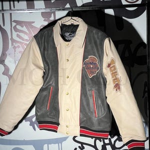 May include: A varsity jacket in cream, dark grey, and black, with red and black striped cuffs and hem. The jacket features a button-up front and patches with text and graphics. The sleeves are cream-colored.