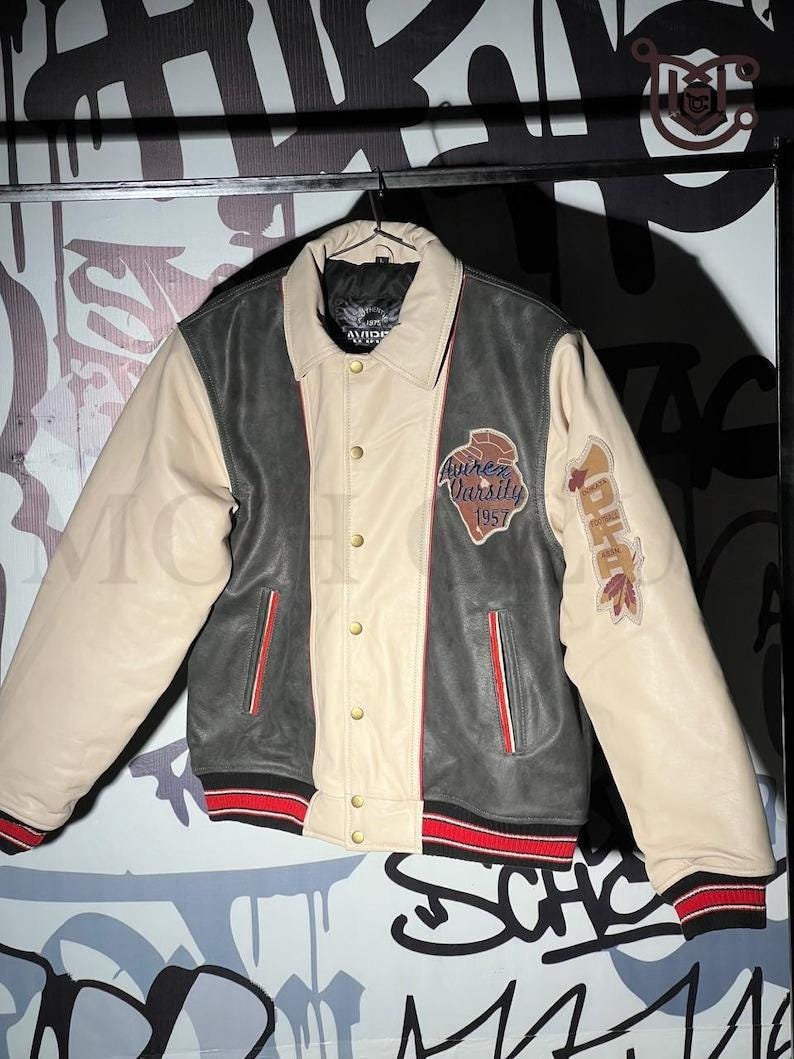 May include: A varsity jacket in cream, dark grey, and black, with red and black striped cuffs and hem. The jacket features a button-up front and patches with text and graphics. The sleeves are cream-colored.