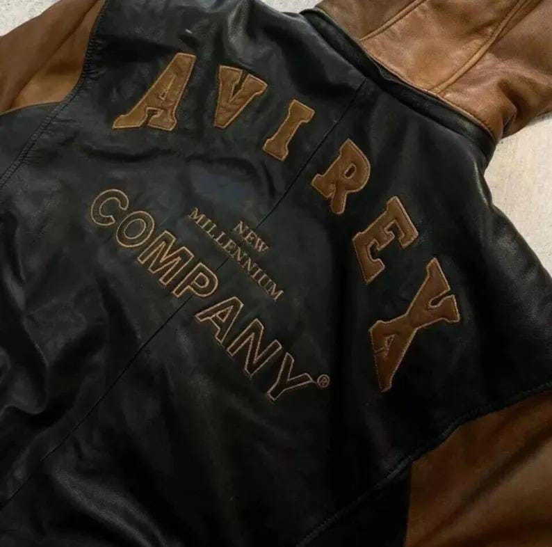 Puede incluir: Una chaqueta de cuero negro con mangas y cuello de cuero marr&oacute;n. La parte posterior presenta el texto AVIREX COMPANY NEW MILLENNIUM en grandes letras de cuero marr&oacute;n aplicadas y cosidas sobre el panel de cuero negro de la prenda.