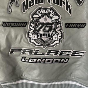May include: A gray leather jacket with white and black embroidered patches. The jacket features the words "New York", "London", and "Tokyo". A central patch reads "75 PALACE LONDON" with the Avirex logo.