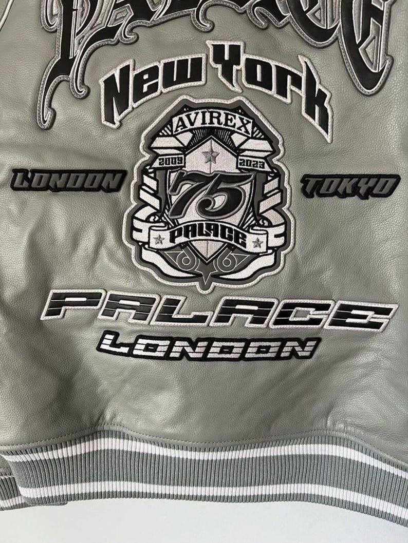 May include: A gray leather jacket with white and black embroidered patches. The jacket features the words "New York", "London", and "Tokyo". A central patch reads "75 PALACE LONDON" with the Avirex logo.