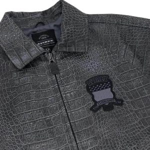 Pode incluir: Casaco cinzento texturizado com padr&atilde;o de pele de crocodilo. O casaco tem fecho de correr e um pequeno patch com o nome da marca e o texto "ESTABLISHED". O casaco tem gola e forro cinzento escuro.
