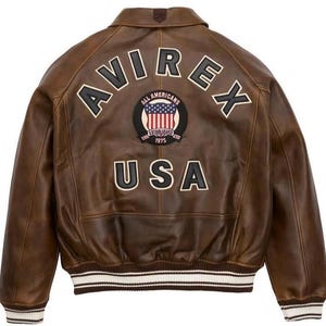May include: Brown leather bomber jacket with the text "AVIREX" and "USA" on the back. Features a patch with the American flag and the words "ALL AMERICANS ESTABLISHED 1975". The jacket has white and brown striped cuffs and hem.