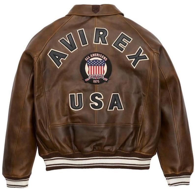 May include: Brown leather bomber jacket with the text "AVIREX" and "USA" on the back. Features a patch with the American flag and the words "ALL AMERICANS ESTABLISHED 1975". The jacket has white and brown striped cuffs and hem.