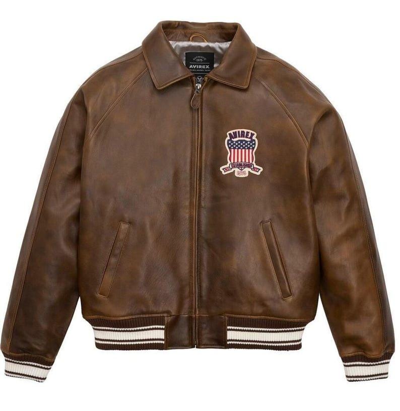 May include: Brown leather jacket with a zip-up front, collar, and ribbed cuffs and hem. Features a patch with an American flag design and the brand name "AVIREX". A classic bomber jacket style.