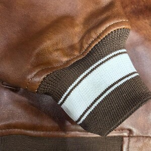 May include: Close-up of a brown leather jacket. The jacket features a ribbed cuff with white stripes. The leather has a smooth texture and a warm, reddish-brown color. The cuff is a dark brown color.
