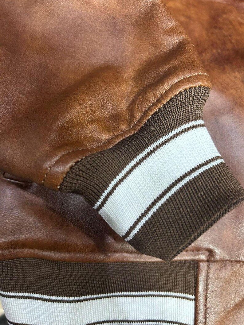 May include: Close-up of a brown leather jacket. The jacket features a ribbed cuff with white stripes. The leather has a smooth texture and a warm, reddish-brown color. The cuff is a dark brown color.