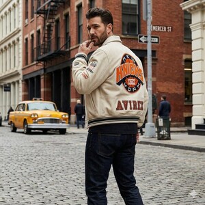May include: Cream-colored varsity jacket with the words "NEW YORK KNICKS" in orange and black lettering on the back. The jacket also features the brand name "AVIREX" in orange lettering. The jacket is worn with dark blue jeans.
