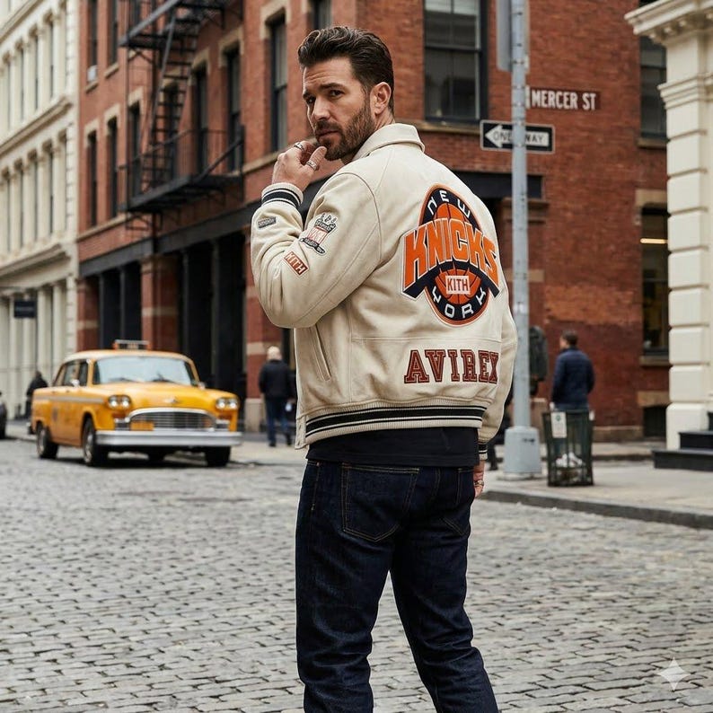 May include: Cream-colored varsity jacket with the words "NEW YORK KNICKS" in orange and black lettering on the back. The jacket also features the brand name "AVIREX" in orange lettering. The jacket is worn with dark blue jeans.