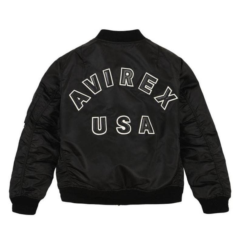 May include: Black bomber jacket with the text "AVIREX USA" in white block letters across the back. The jacket has a ribbed collar, cuffs, and hem. It features a zippered pocket on the left sleeve.