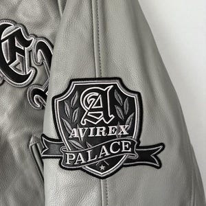 May include: Close-up of a gray leather jacket with black and white embroidered patches. The patches feature the text "AVIREX PALACE" and decorative lettering. The jacket has a classic varsity style.