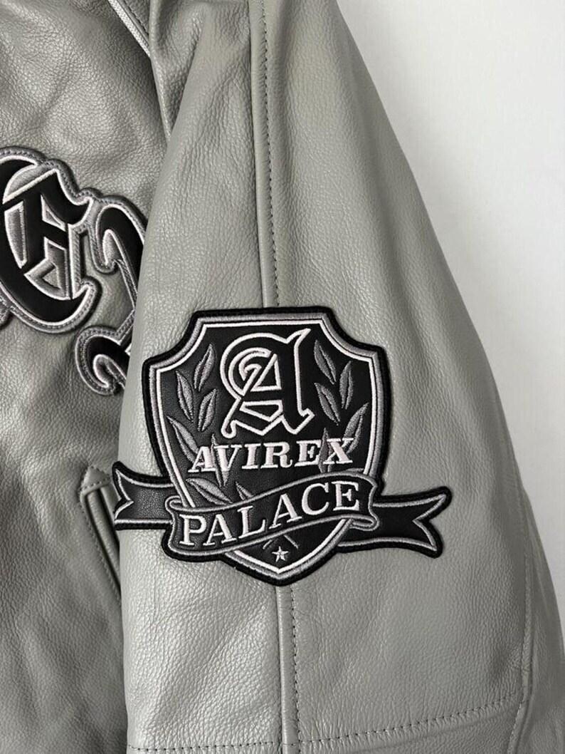 May include: Close-up of a gray leather jacket with black and white embroidered patches. The patches feature the text "AVIREX PALACE" and decorative lettering. The jacket has a classic varsity style.