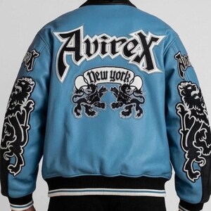 Puede incluir: Chaqueta de cuero azul claro con detalles en negro y blanco. La parte trasera presenta la palabra "Avirex" en letras blancas grandes, con "New York" debajo. Gr&aacute;ficos de leones negros en las mangas y la espalda.
