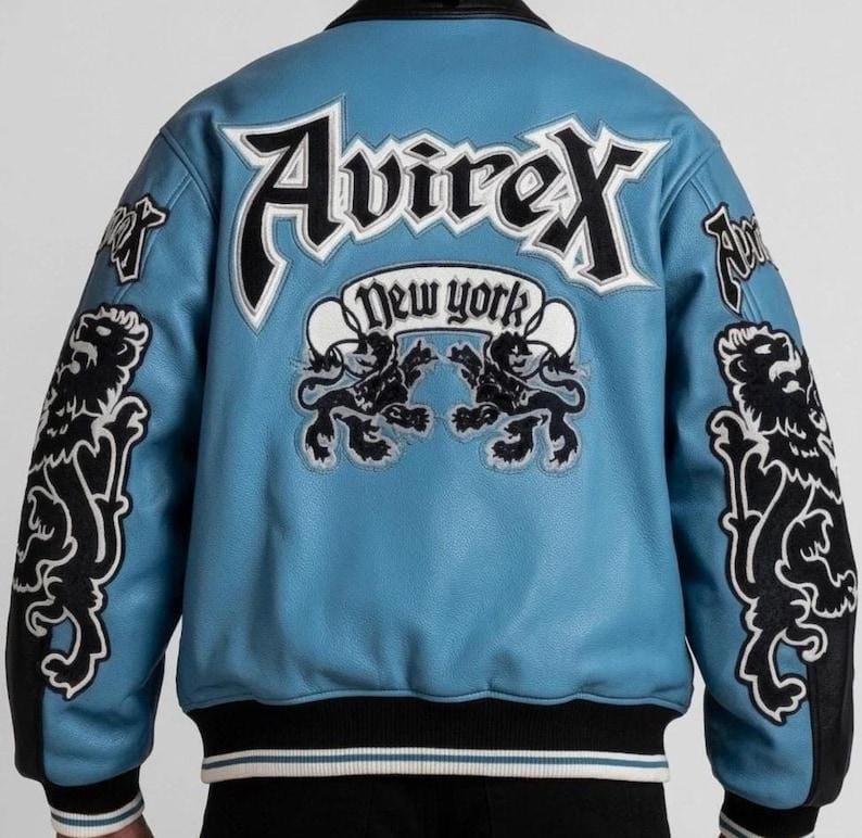 Puede incluir: Chaqueta de cuero azul claro con detalles en negro y blanco. La parte trasera presenta la palabra "Avirex" en letras blancas grandes, con "New York" debajo. Gr&aacute;ficos de leones negros en las mangas y la espalda.